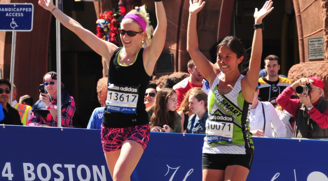 Two runners crossing the Boston Marathon finish line with arms raised, one wearing a RunSmart singlet